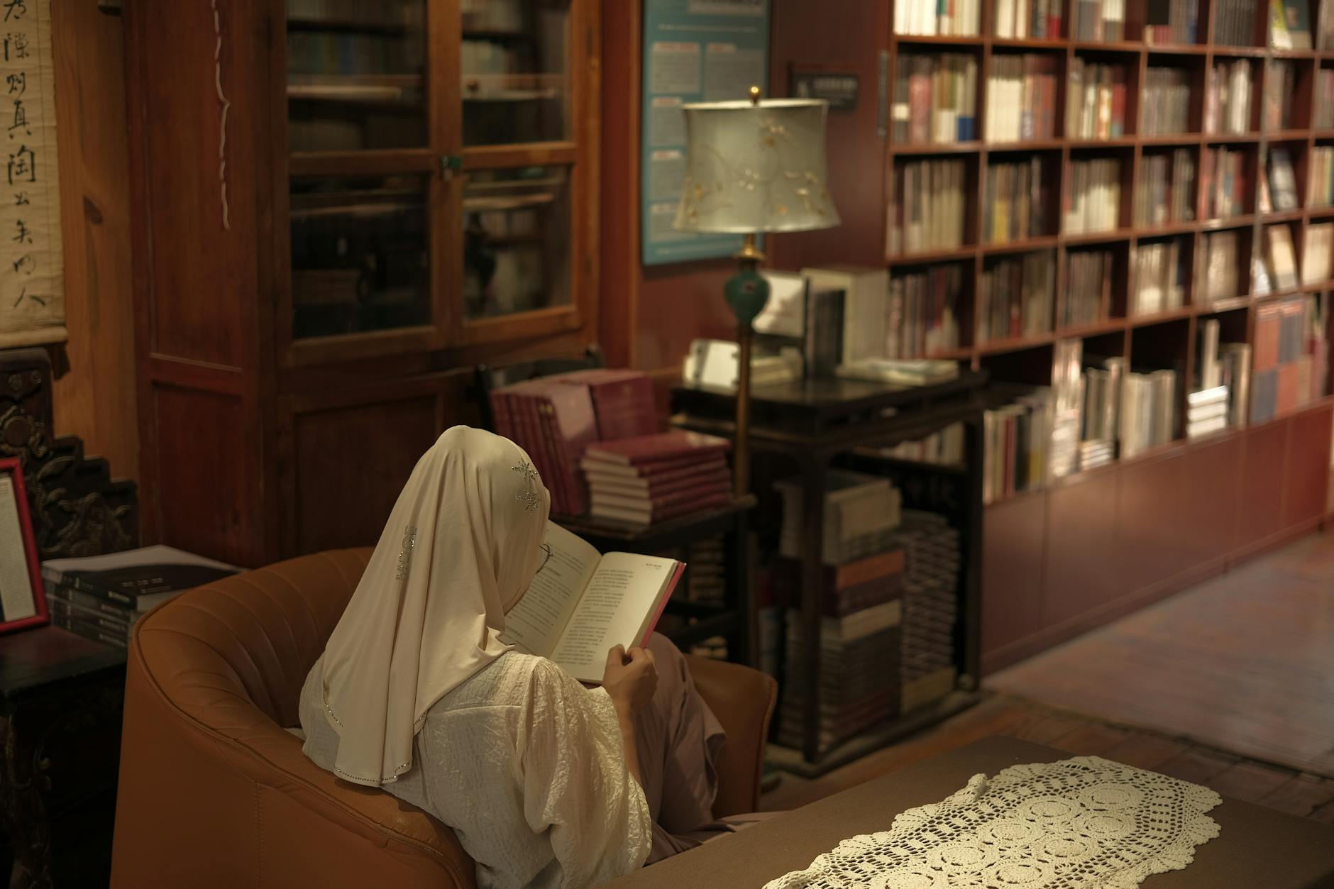 A woman in a cozy library setting is seen reading a book while wearing a hijab.