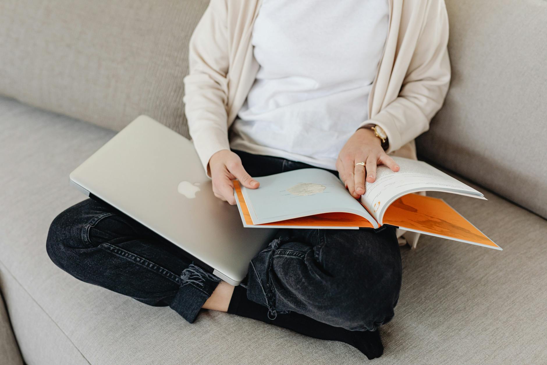 An adult sitting on a sofa, reading a book with a laptop nearby, conveying relaxation and leisure.