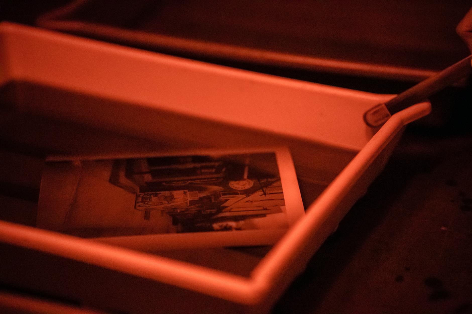 Monochrome photo in a developing tray with red light, showcasing darkroom process.